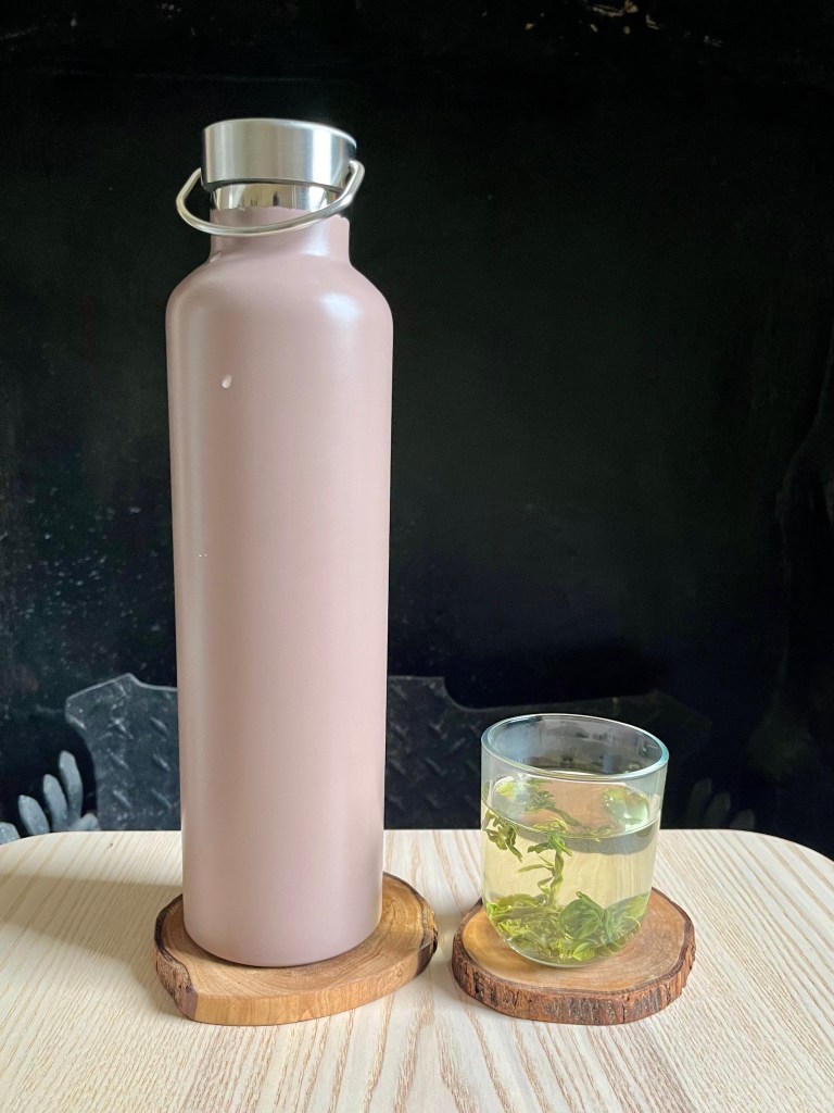 Liter thermos and a glass with green tea infusing loose in it on a wooden table in front of a dark background