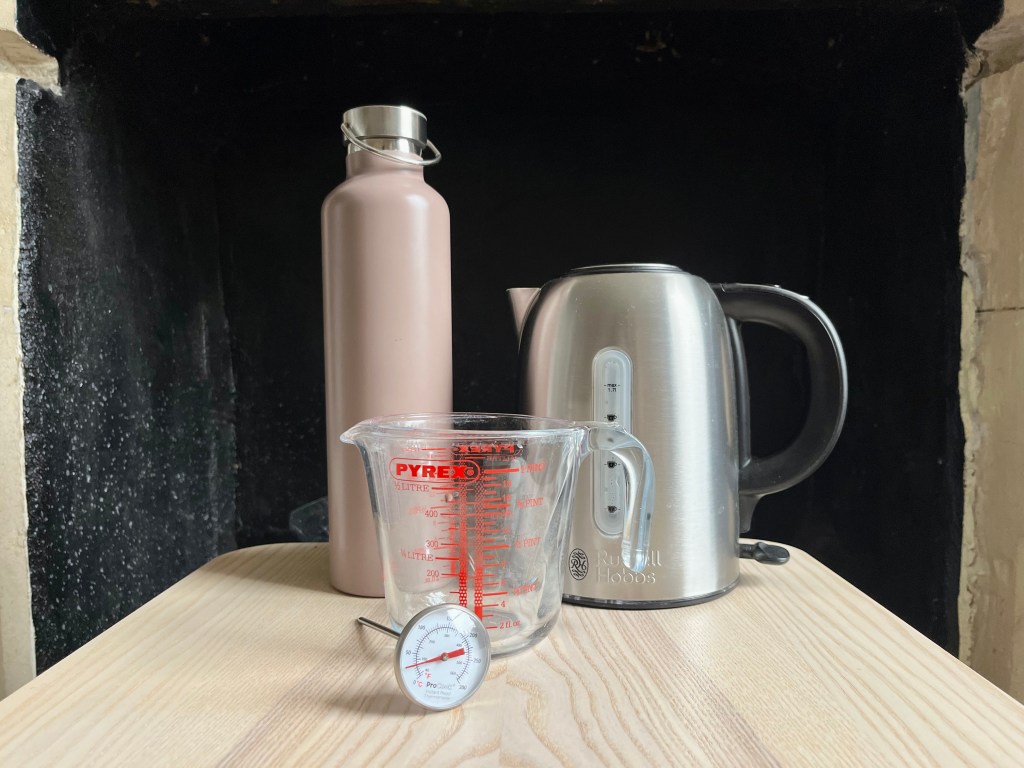 A thermos, kettle, glass jug, and instant read thermometer on a wooden table in front of a dark background