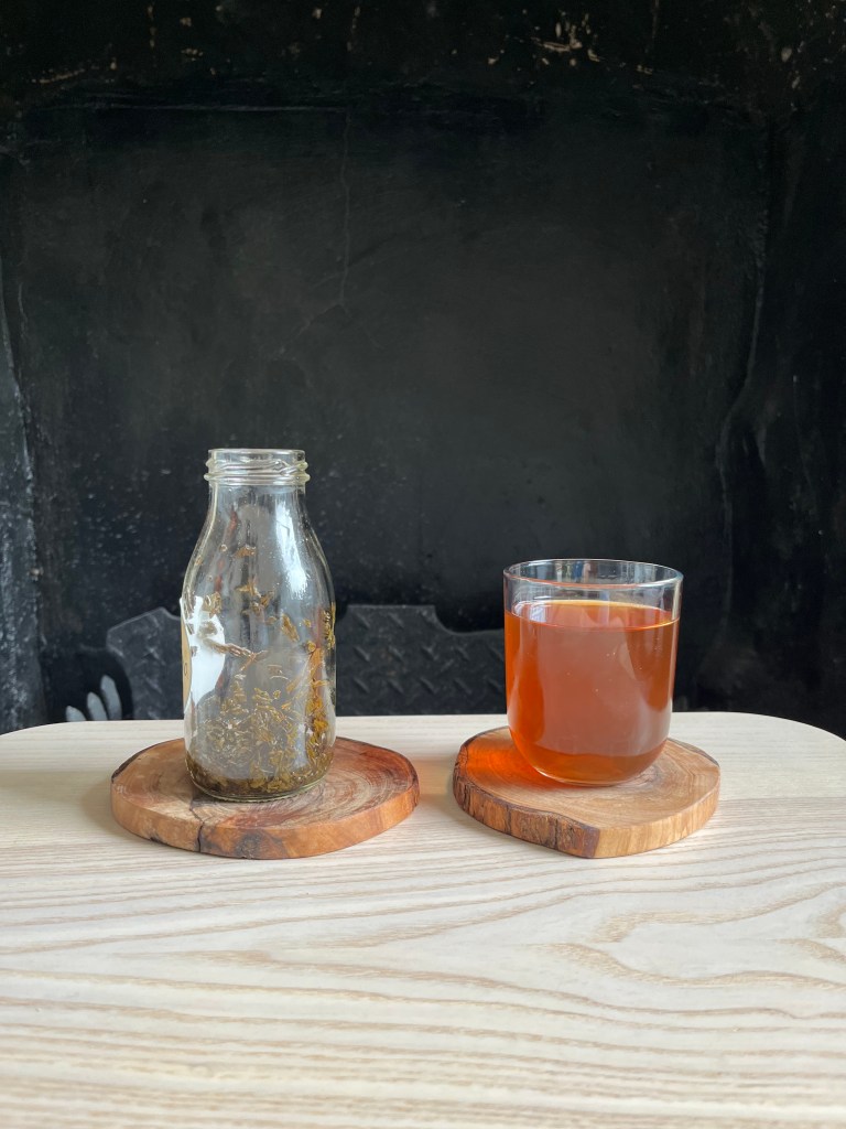 Glass cold-brew bottle with tea leaves in it next to glass cup full of dark rose-red cold-brewed tea