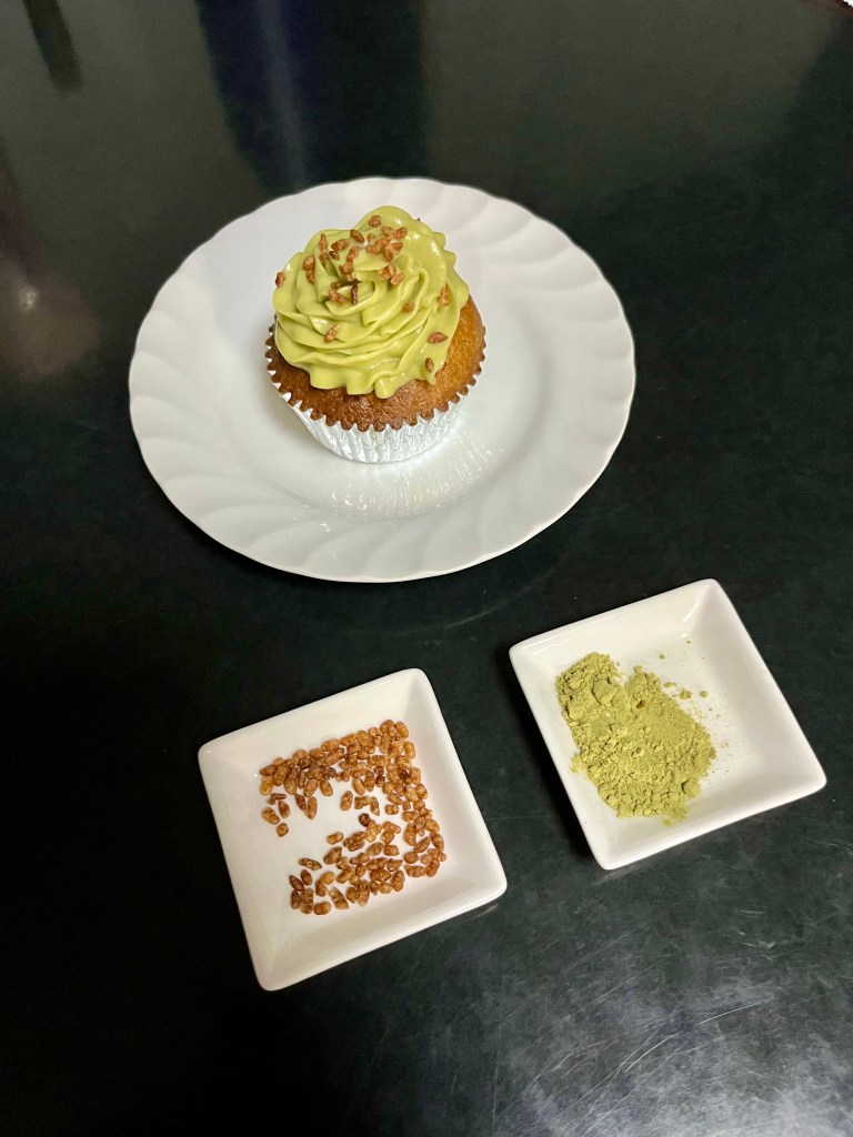 One finished cupcake on a white plate, with trays of genmai and genmaicha powder