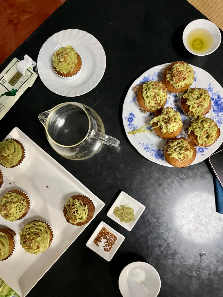 A table with multiple plates of cupcakes, some teacups, pitcher, and trays of genmai and genmaicha powder