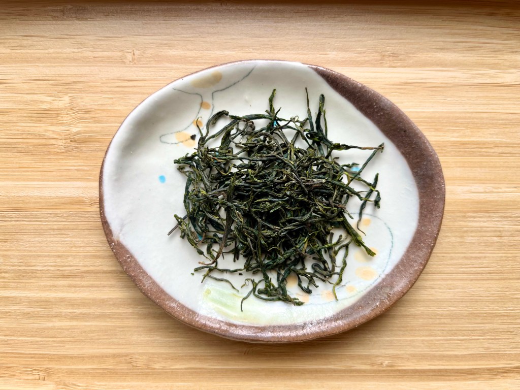 A tray of dry tea leaves, dark rich green with some brighter highlights, some quite curly and some straighter but all quite thinly rolled