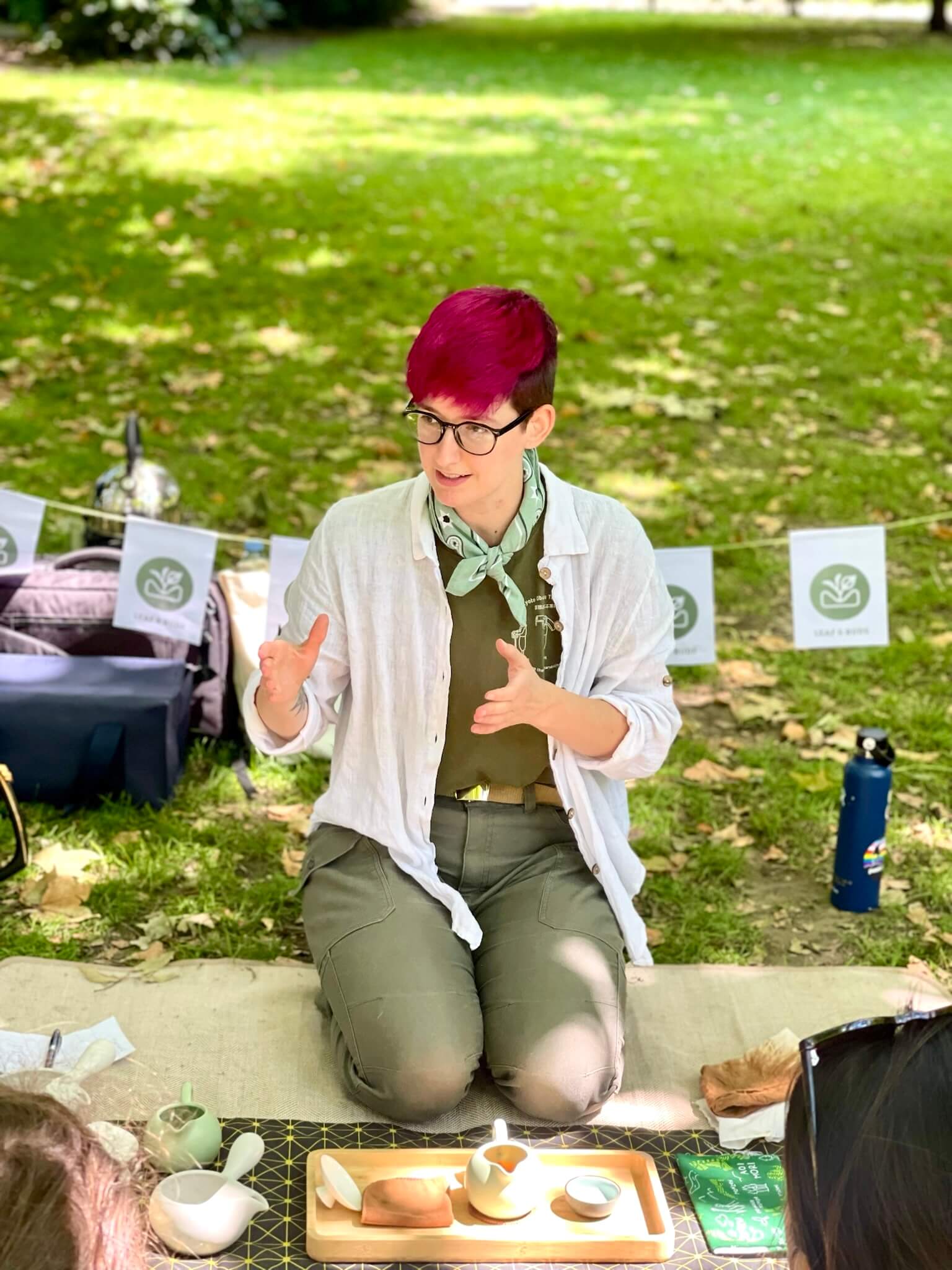 Maren sits in seiza on a picnic blanket on the ground behind a tray of teaware, explaining tea processing
